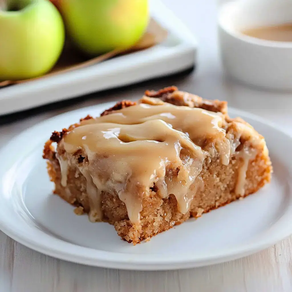 A slice of moist apple cake topped with a creamy caramel glaze, served on a white plate with green apples in the background.