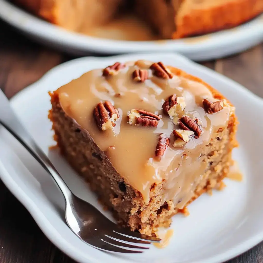 A slice of cake topped with a caramel glaze and chopped pecans on a white plate, with a fork resting beside it.