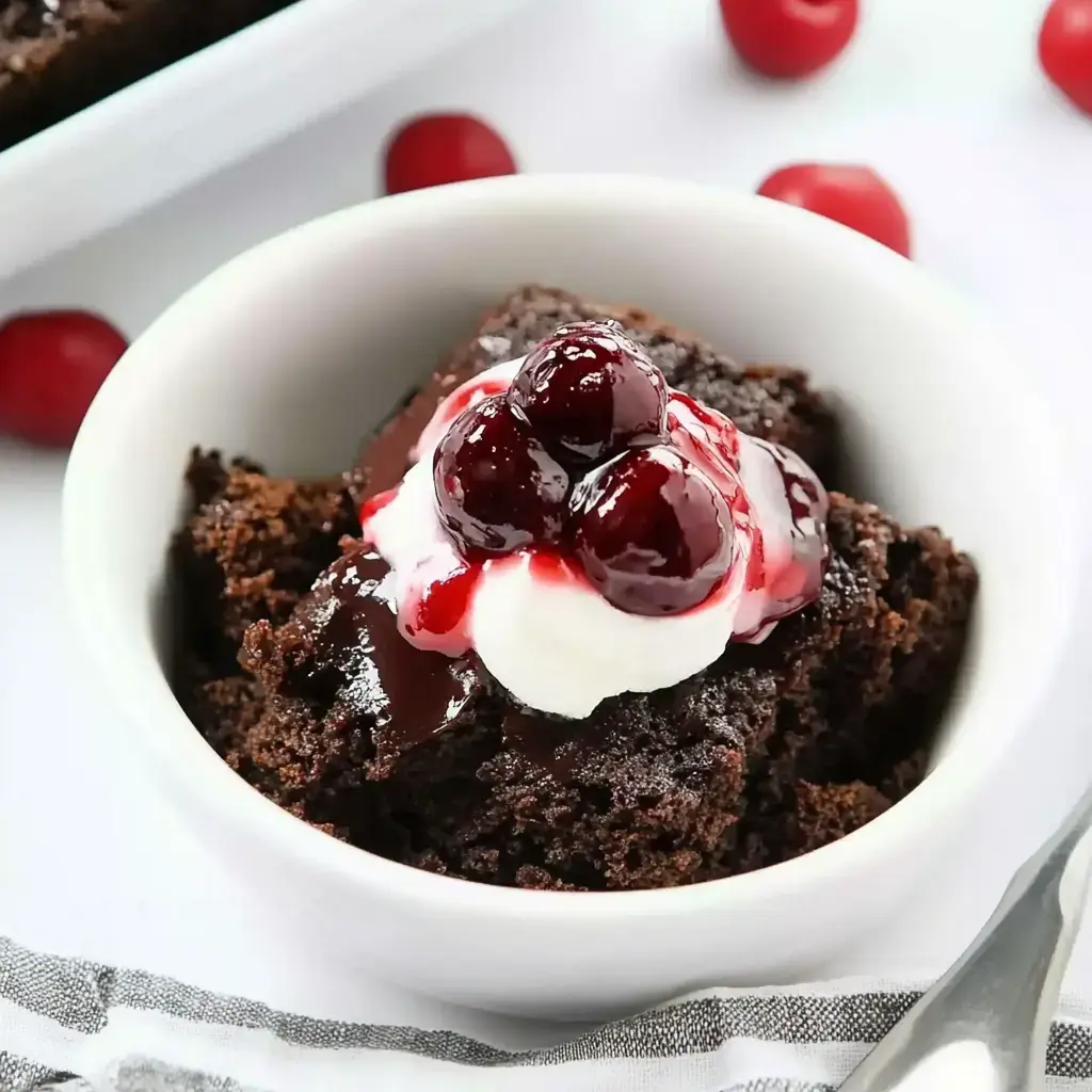 A bowl of chocolate cake topped with whipped cream and cherry sauce, surrounded by scattered cherries.