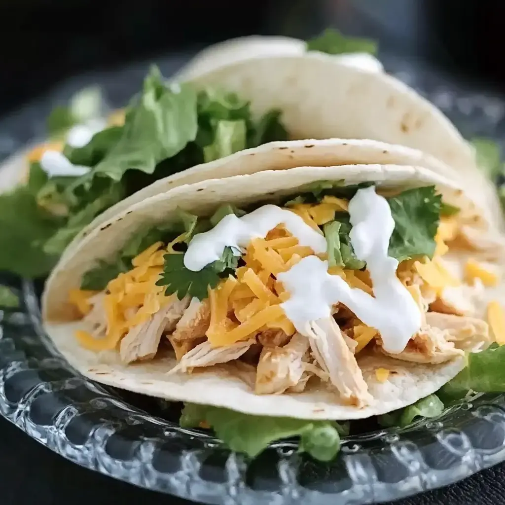 Three soft tacos filled with shredded chicken, shredded cheese, lettuce, and topped with sour cream and cilantro, served on a decorative plate.