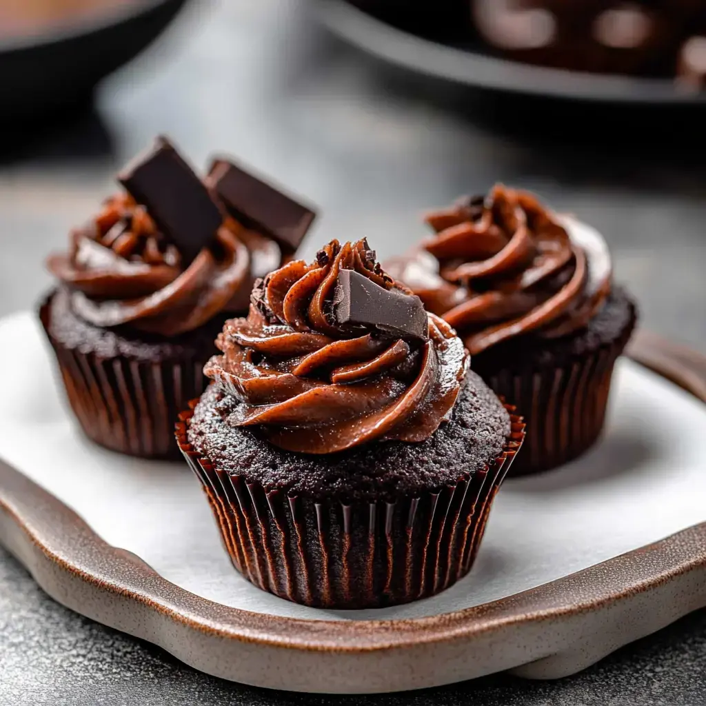 Three chocolate cupcakes with swirls of chocolate frosting and topped with pieces of dark chocolate.