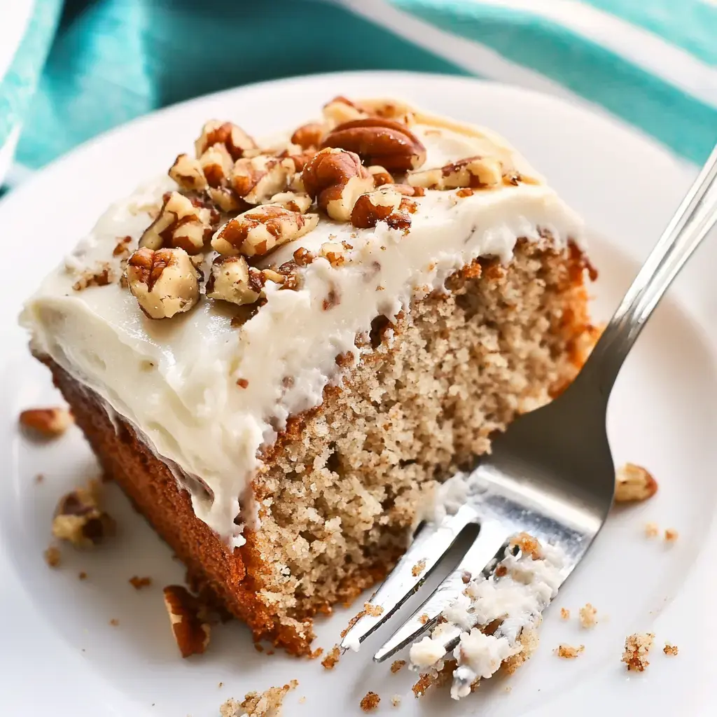 A slice of moist cake topped with creamy frosting and chopped nuts sits on a white plate, with a fork beside it.