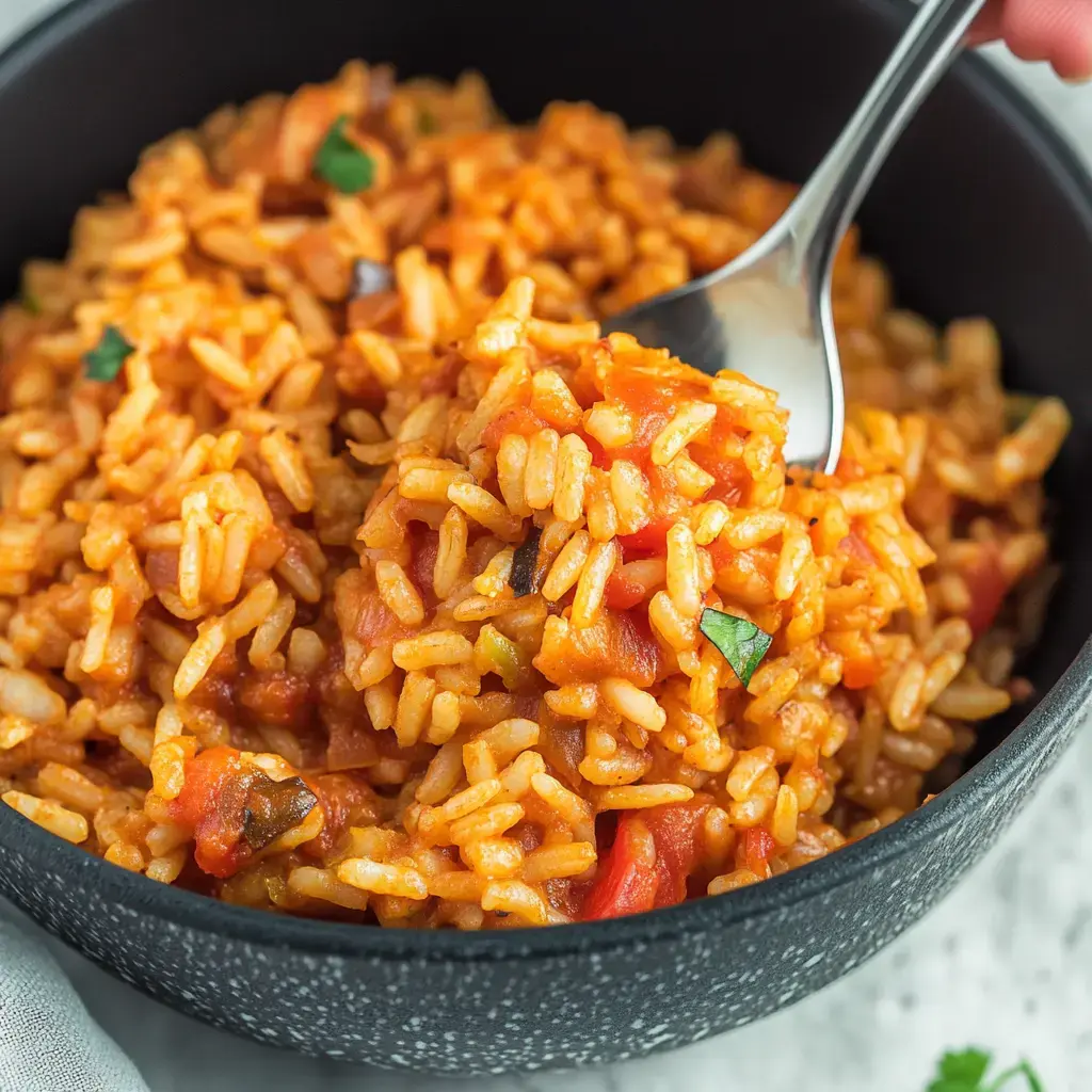 A close-up of a bowl of colorful, seasoned rice mixed with tomatoes and herbs, with a spoon prominently lifting some from the bowl.