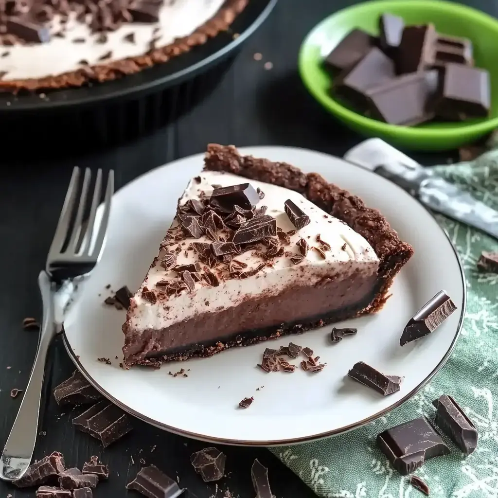 A slice of chocolate cream pie sits on a white plate, topped with chocolate shavings, alongside a fork and pieces of chocolate in the background.