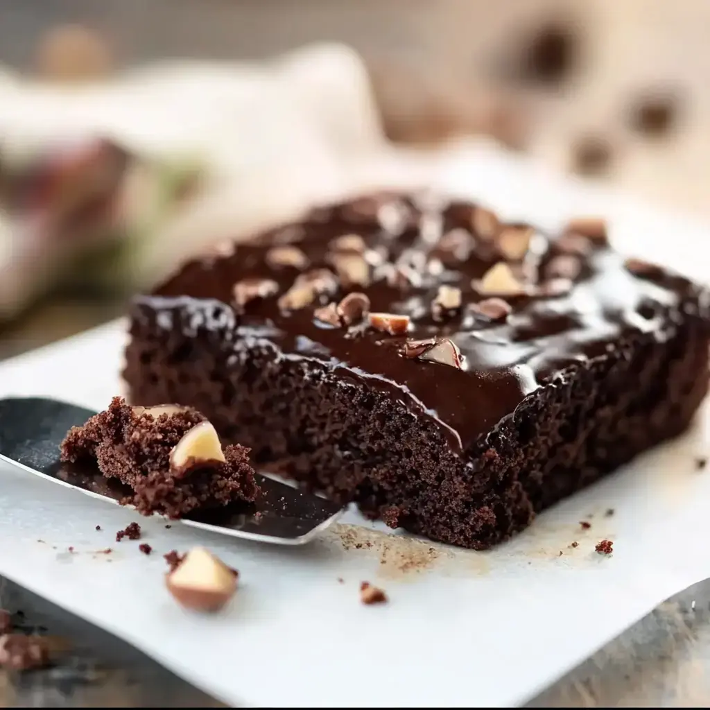 A piece of chocolate cake with glossy frosting and chopped nuts sits on a plate, with a serving utensil holding a bite-sized piece nearby.