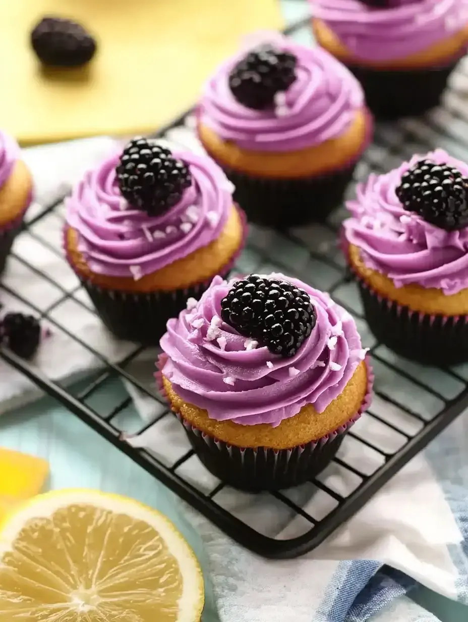 A close-up of decorated cupcakes with purple frosting and blackberries on top, presented on a cooling rack next to lemon slices.