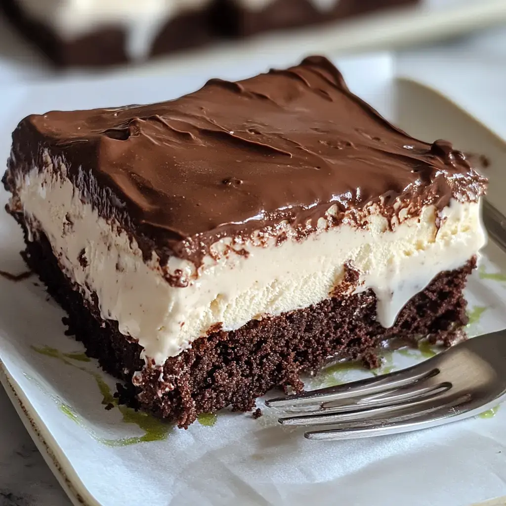 A slice of chocolate-covered ice cream cake with layers of brownie and vanilla ice cream sits on a white plate next to a fork.