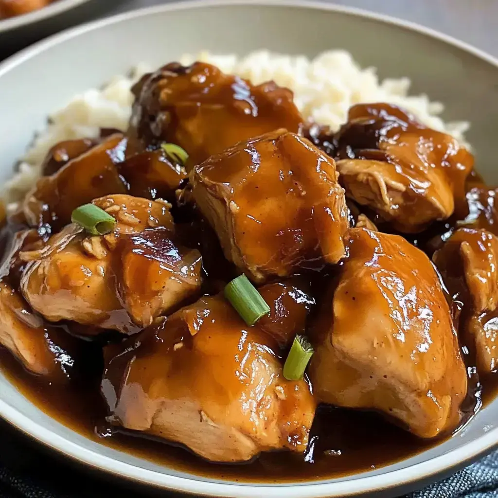 A close-up of tender chicken pieces coated in a glossy brown sauce served over a bed of white rice, garnished with green onions.