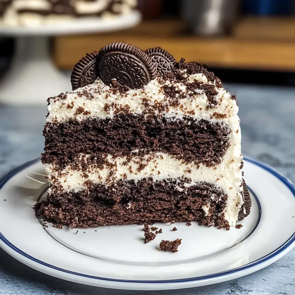 A slice of chocolate cake layered with white frosting and topped with crushed Oreos and whole Oreo cookies, presented on a white plate.