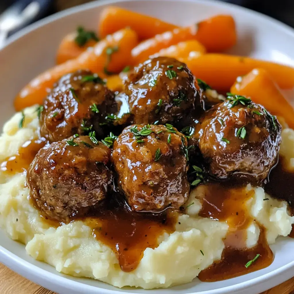 A plate of meatballs covered in brown gravy is served on a bed of creamy mashed potatoes, accompanied by orange carrots.