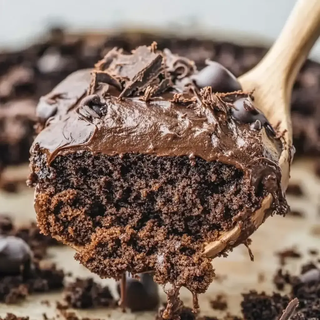 A piece of moist chocolate cake with rich chocolate frosting is being lifted by a wooden spoon, surrounded by crumbled cake and chocolate chips.