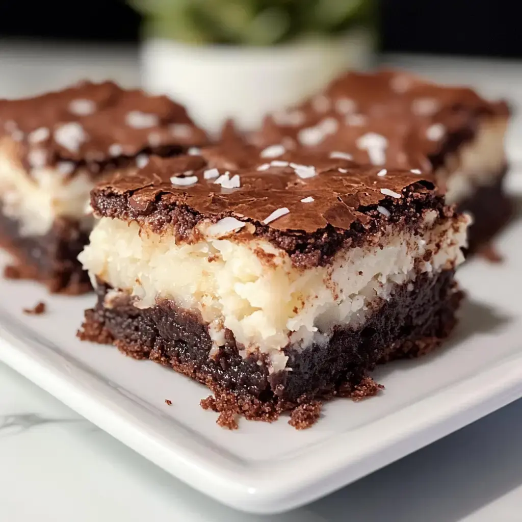 A close-up of a plate featuring decadent layered dessert bars with a chocolate top, coconut filling, and a dark brownie base.