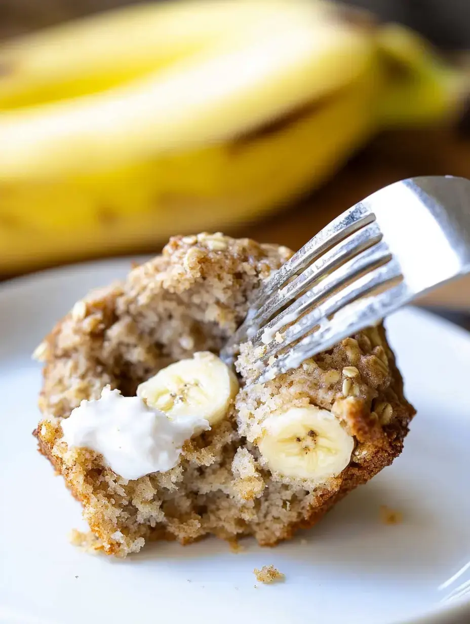 A partially eaten banana muffin with visible banana slices and a dollop of cream is on a plate, with a banana in the background.