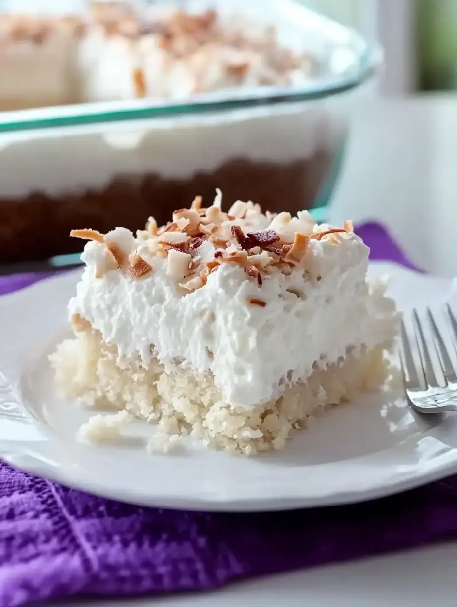 A slice of coconut cream dessert with whipped cream and toasted coconut flakes served on a white plate, accompanied by a silver fork, on a purple cloth.