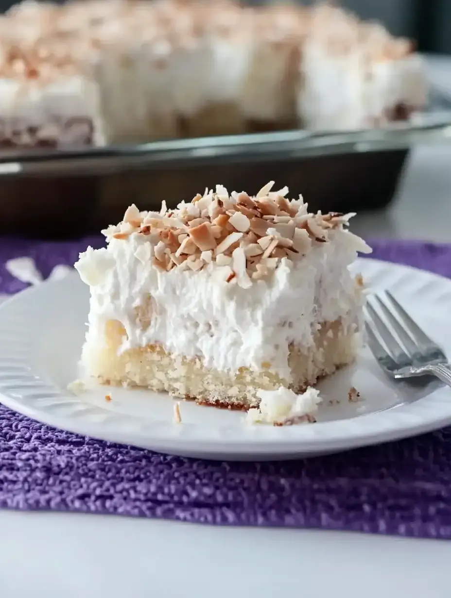 A slice of coconut cream cake topped with shredded coconut sits on a white plate next to a fork, with more cake in the background.