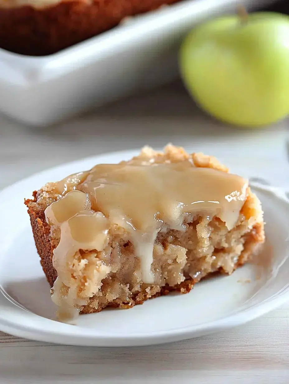 A slice of apple cake topped with a glossy glaze, served on a white plate with a green apple in the background.