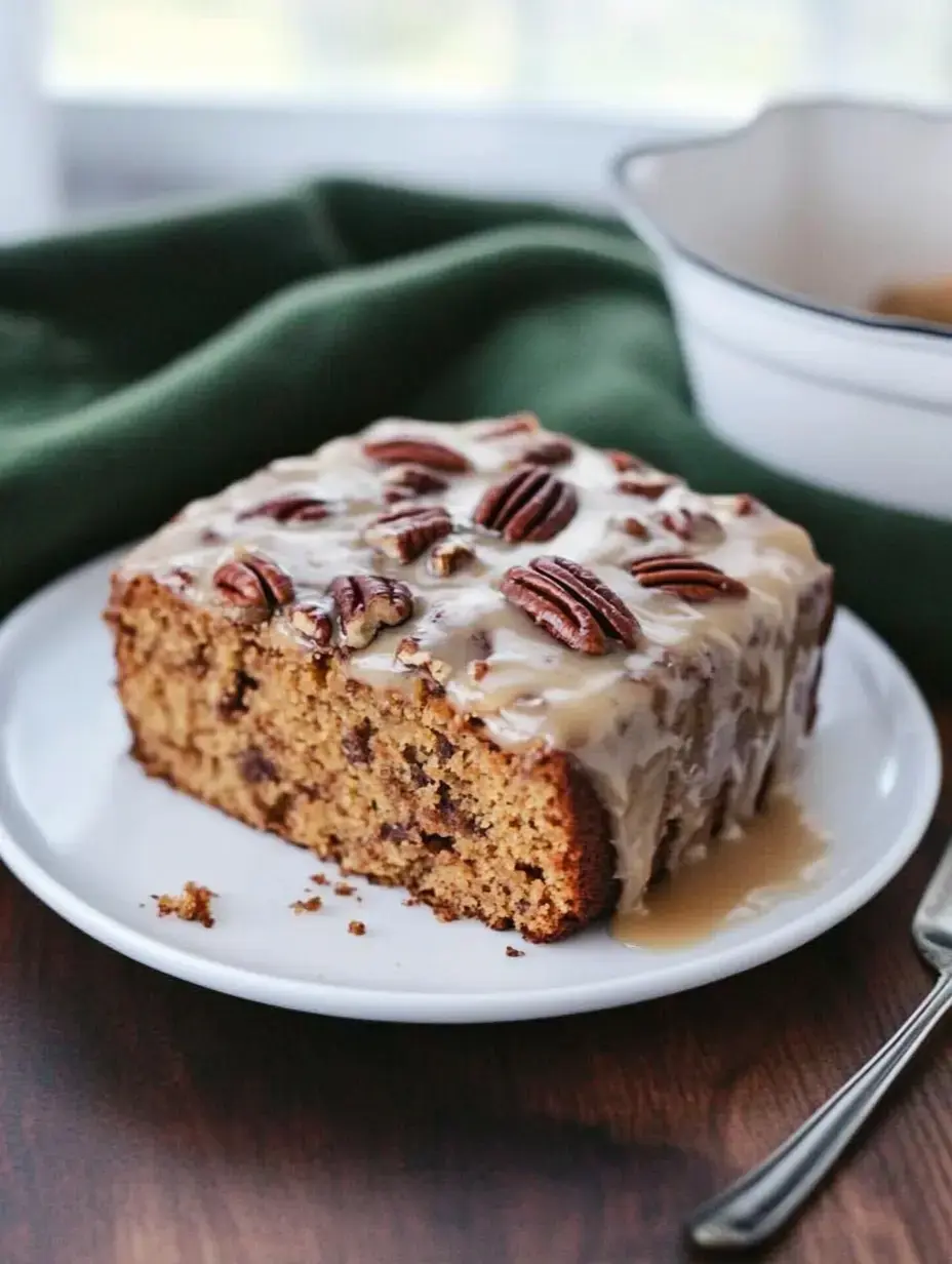 A slice of cake topped with a creamy glaze and pecans, served on a white plate with a green cloth in the background.