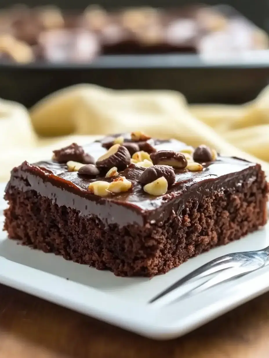 A rich chocolate cake slice topped with chocolate frosting, chocolate chips, and chopped nuts, served on a white plate.