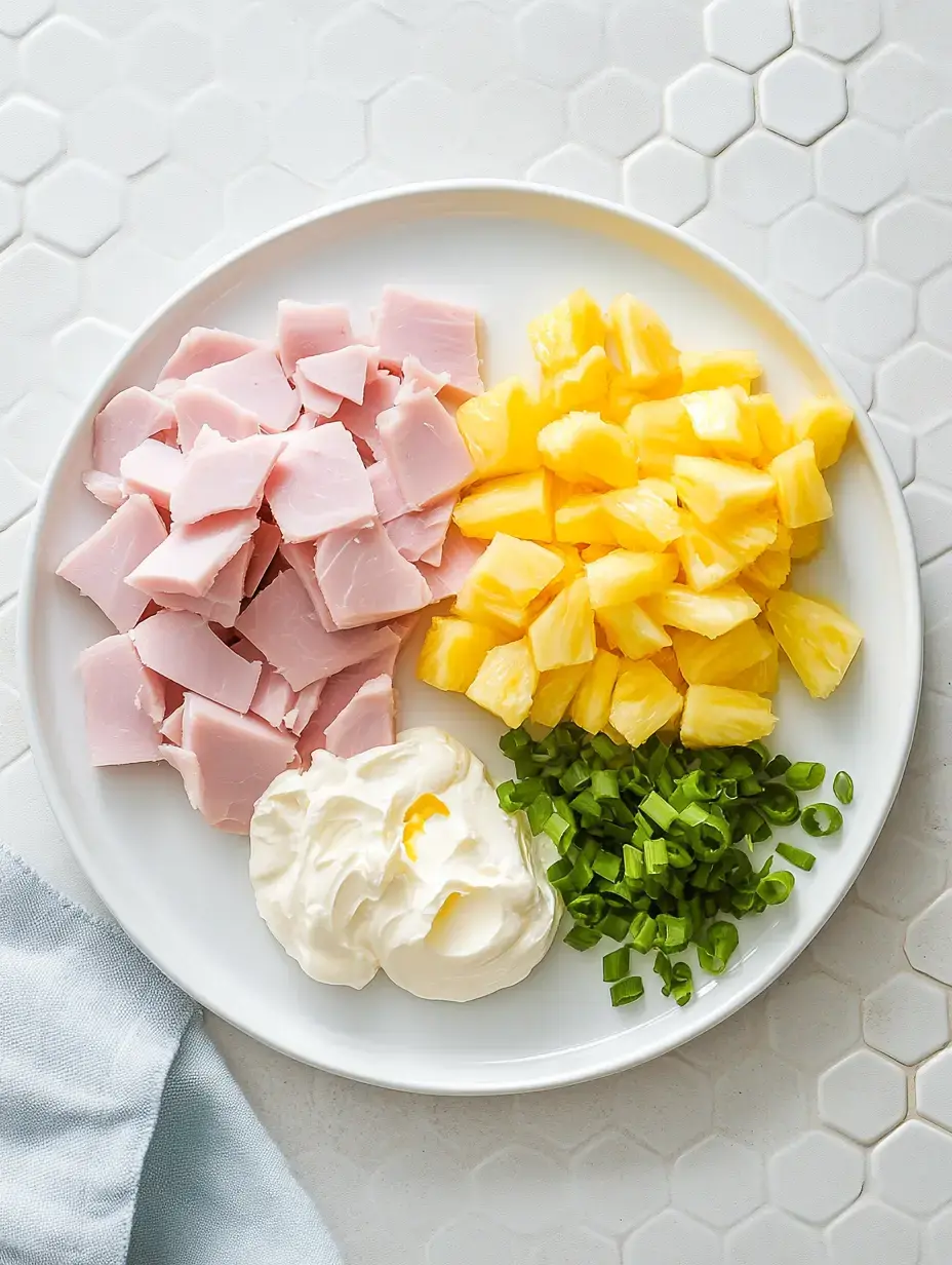 A plate featuring diced ham, pineapple chunks, green onions, and a dollop of cream on a textured surface.