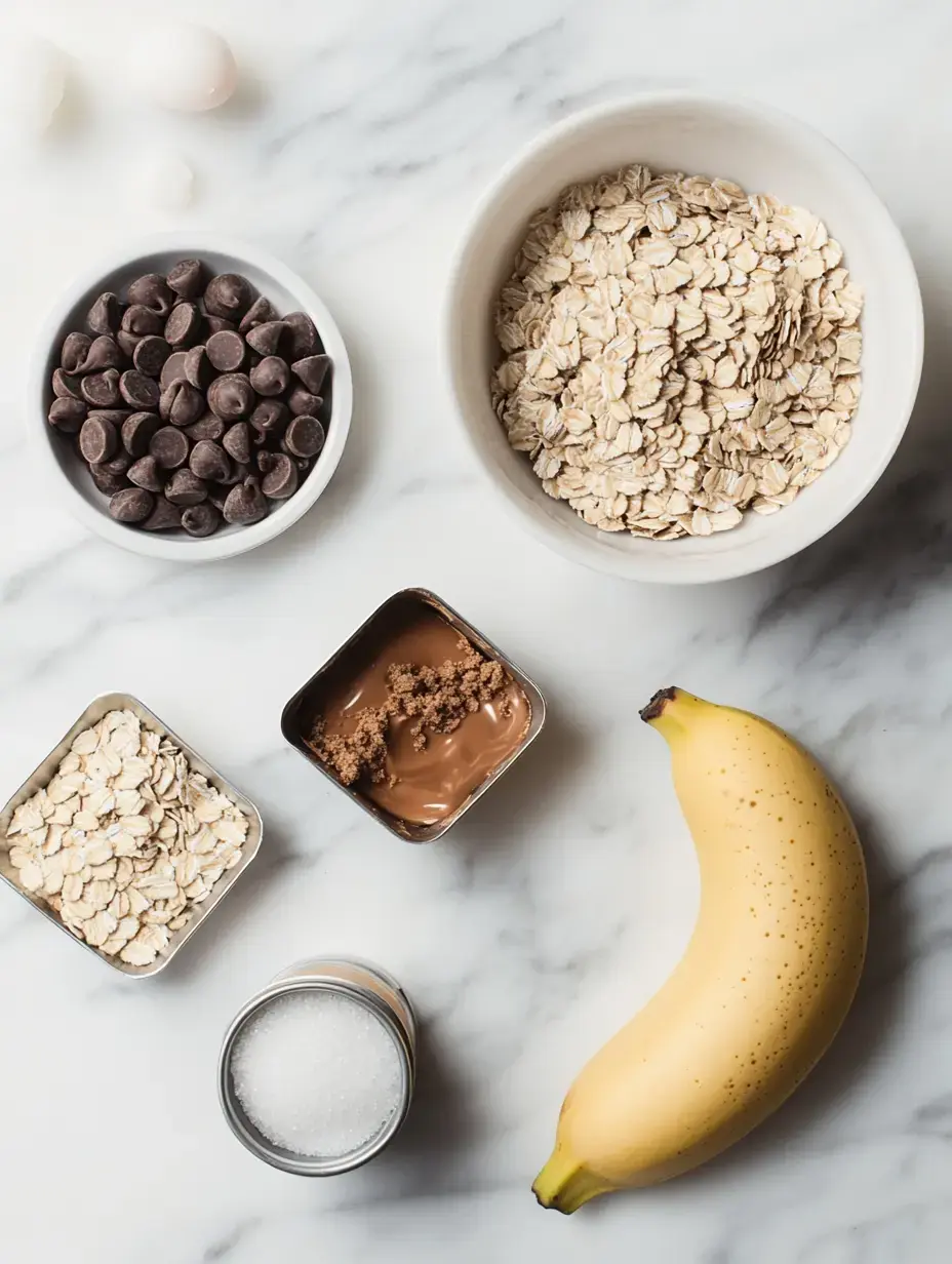 A flat lay of ingredients for a recipe, including oats, chocolate chips, banana, sugar, brown sugar, and eggs on a marble surface.