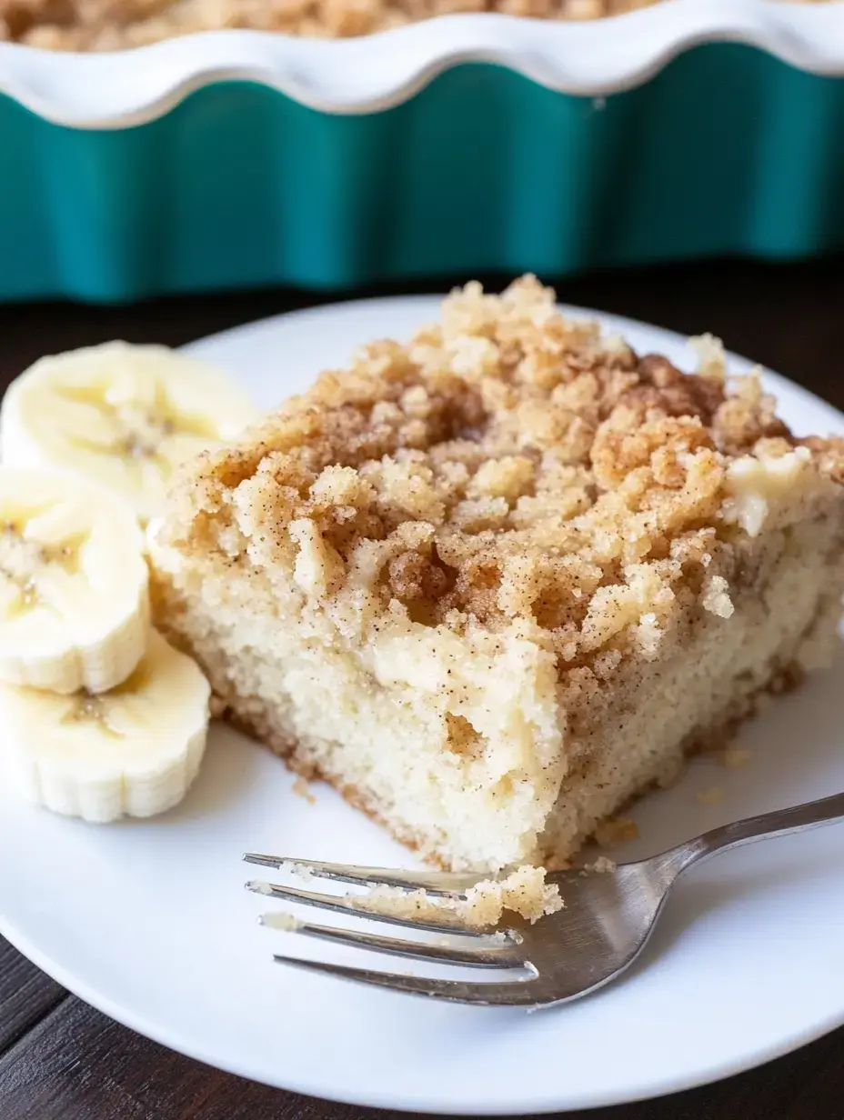 A slice of crumb cake is served on a white plate next to sliced bananas and a fork.