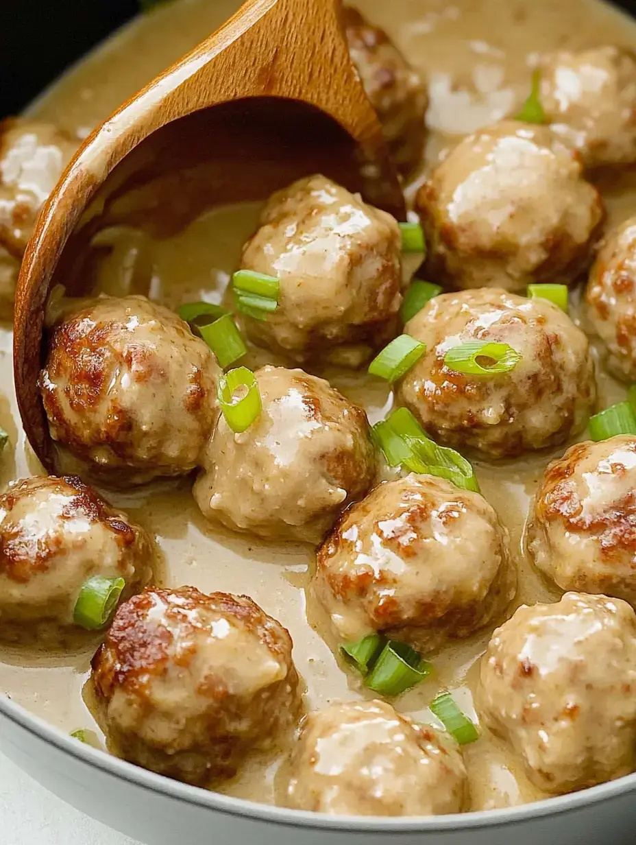 A close-up of meatballs in a creamy sauce, garnished with chopped green onions, with a wooden spoon scooping some from the dish.