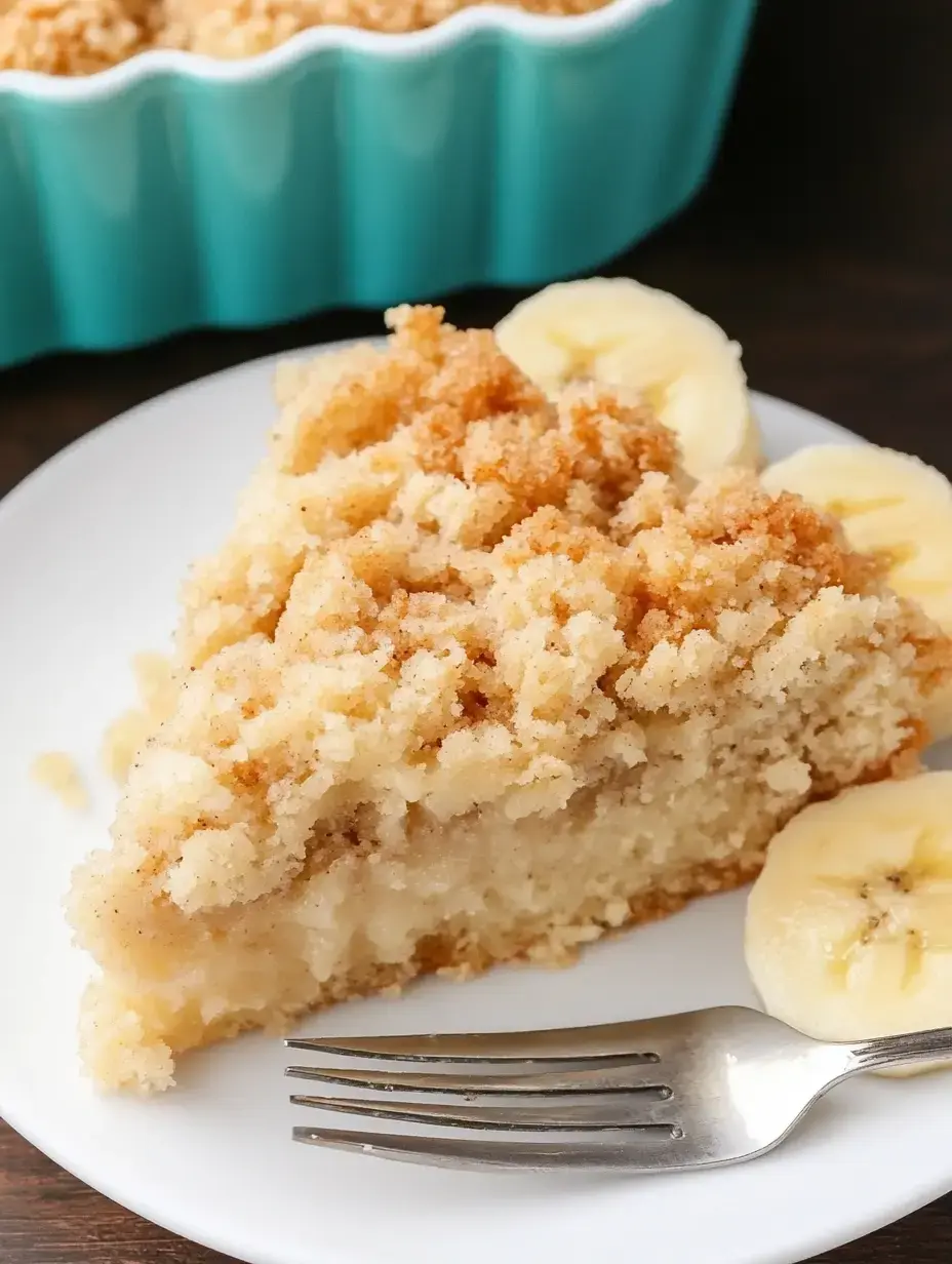 A slice of crumbly banana cake is plated next to banana slices and a fork, with a light blue dish of cake in the background.