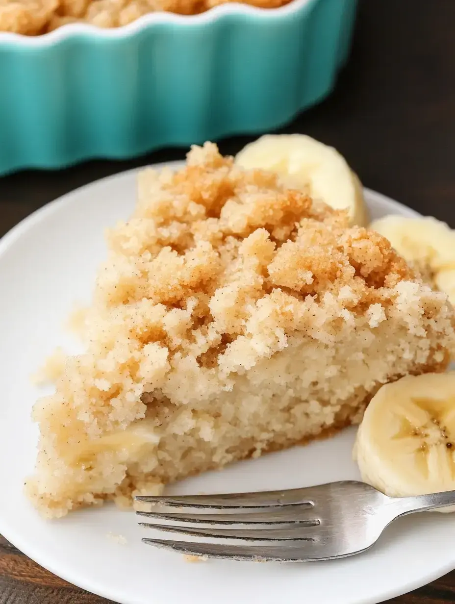 A close-up of a slice of crumbly cake on a white plate, served with banana slices and a fork, with a turquoise baking dish in the background.