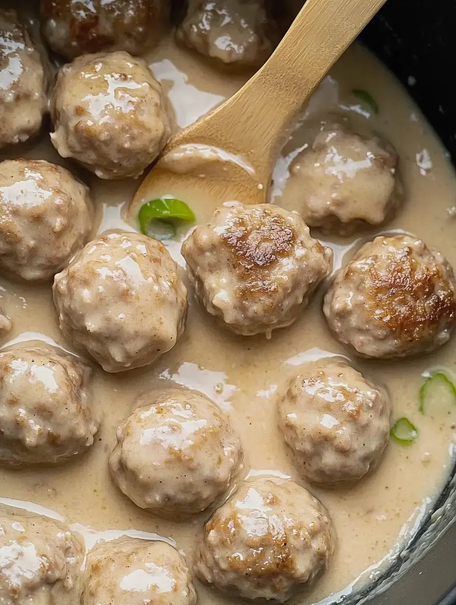 A close-up of meatballs simmering in a creamy sauce, with a wooden spoon resting beside them.