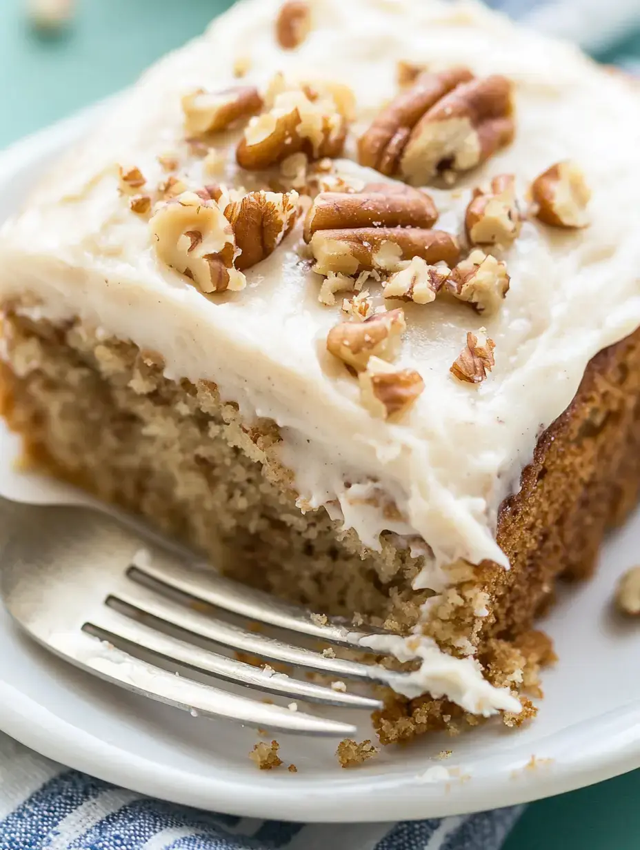 A slice of cake topped with cream frosting and chopped pecans sits on a plate next to a fork.