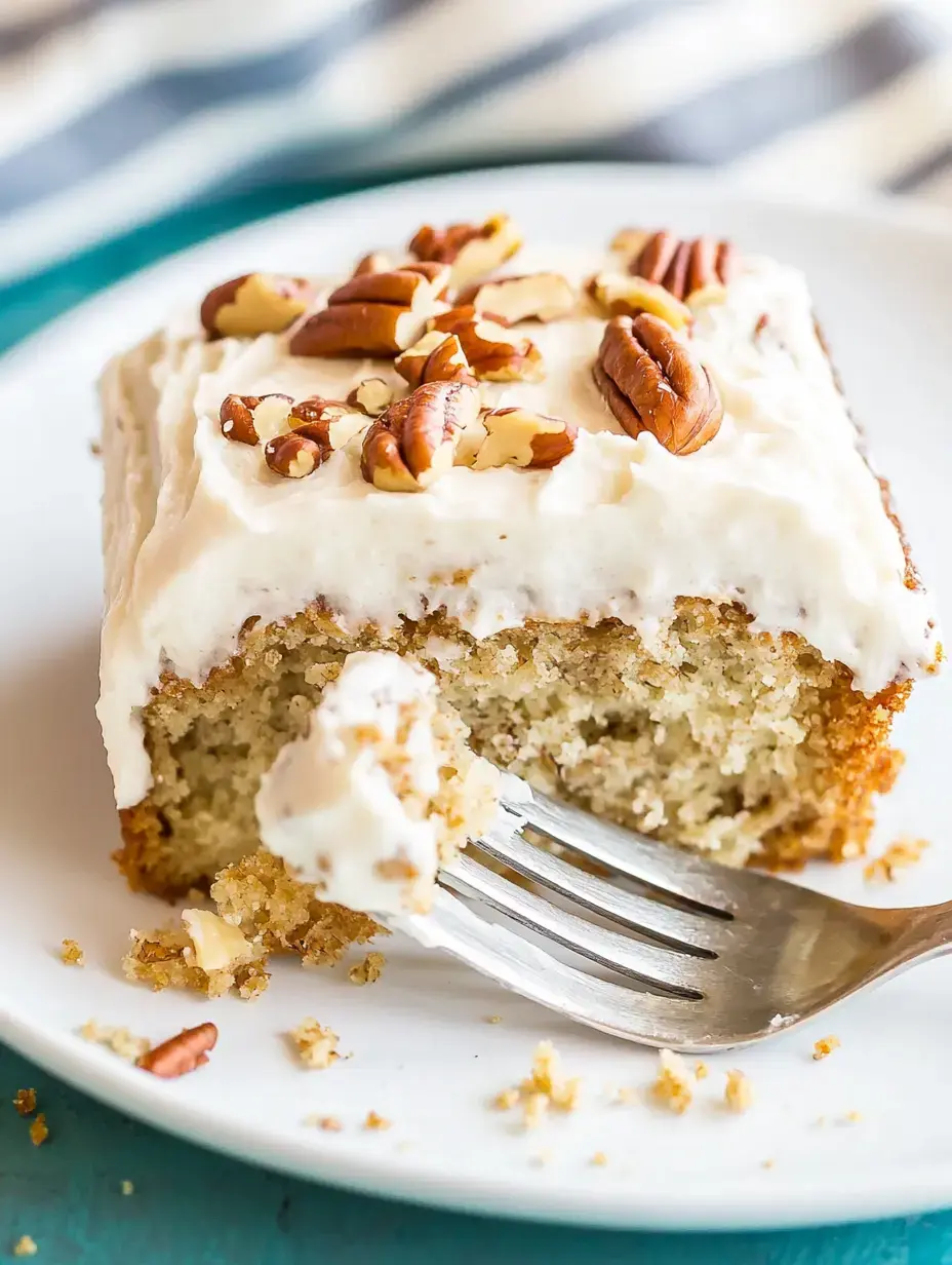 A slice of cake topped with cream frosting and chopped pecans is shown on a white plate, with a fork partially lifting a bite of the cake.