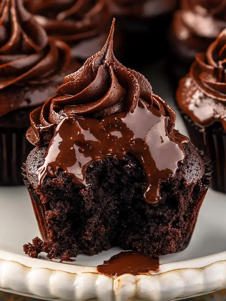 A chocolate cupcake with swirls of chocolate frosting and a glossy chocolate drizzle, partially bitten into, sits on a decorative plate.