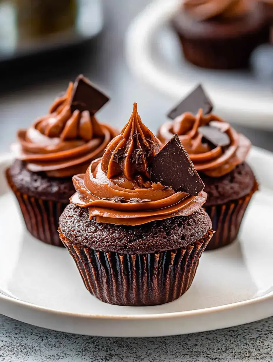 Three chocolate cupcakes with swirls of chocolate frosting and dark chocolate garnishes are displayed on a white plate.