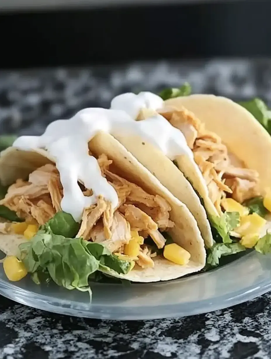 A close-up of three tacos filled with shredded chicken, lettuce, and corn, topped with sour cream on a glass plate.