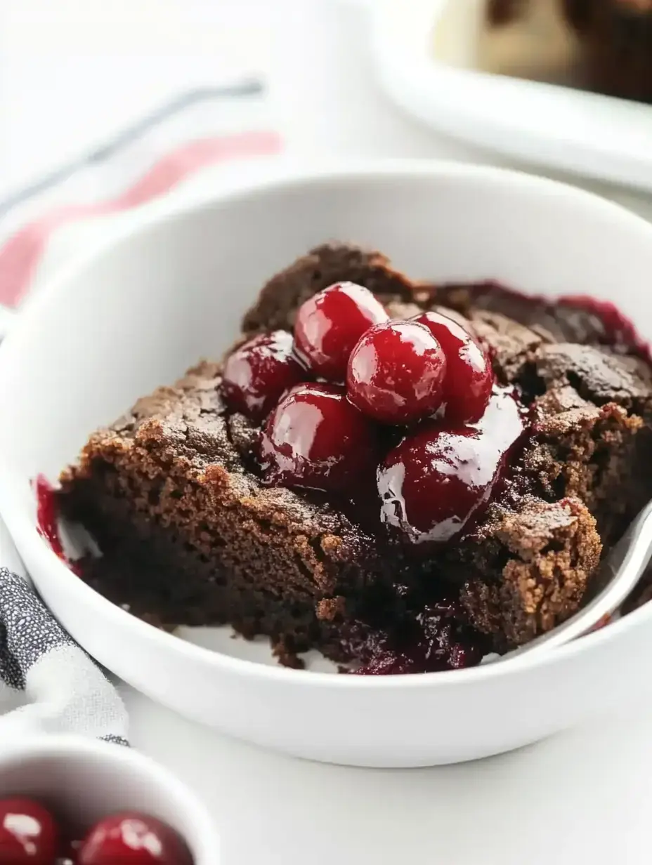 A portion of chocolate cake topped with glossy cherries sits in a white bowl.