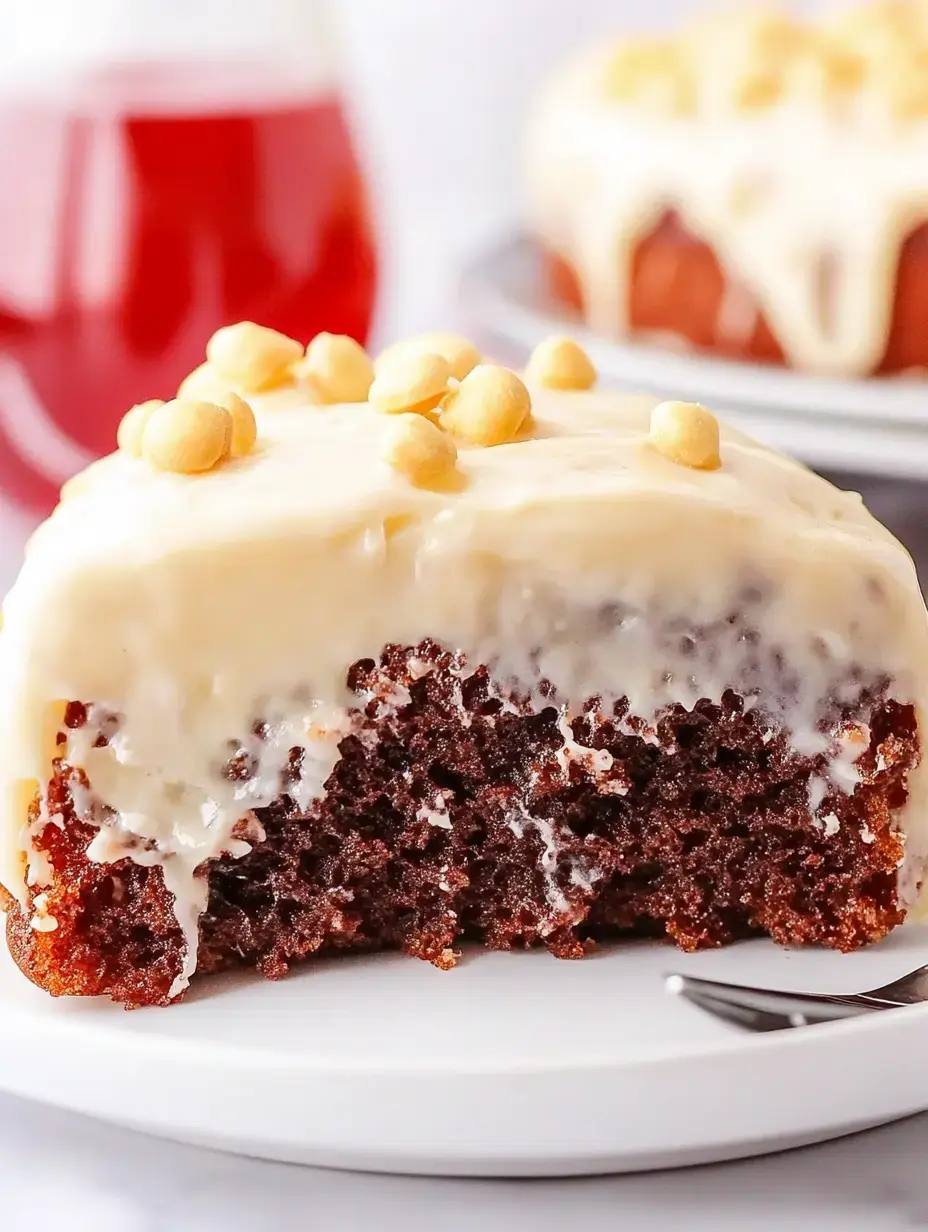 A close-up of a slice of moist chocolate cake topped with creamy frosting and scattered with golden chips, served on a white plate with a glass of red drink in the background.