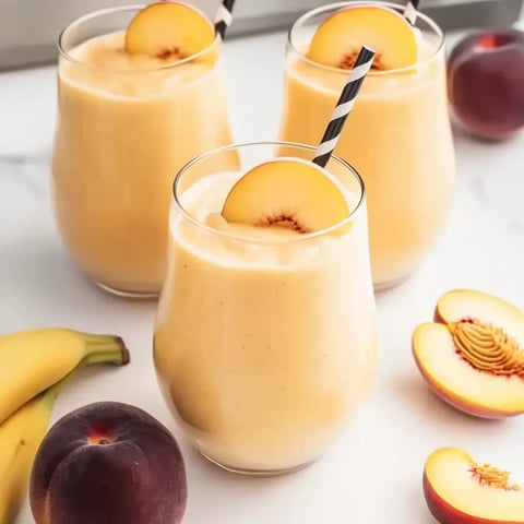 Three creamy peach smoothies topped with peach slices and black-and-white striped straws, with fresh peaches and bananas nearby.