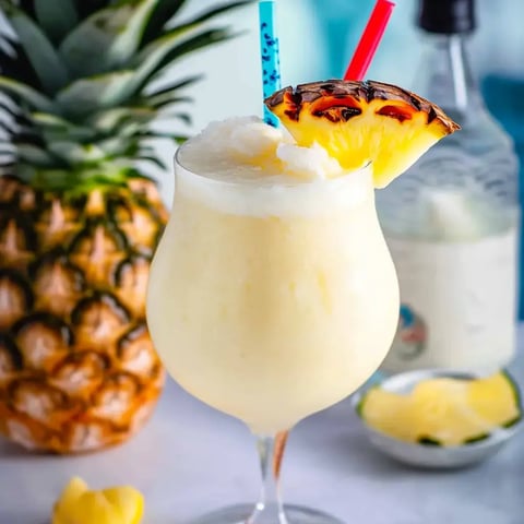 A creamy frozen cocktail with a pineapple slice and colorful straws is displayed beside a fresh pineapple and a bottle in the background.