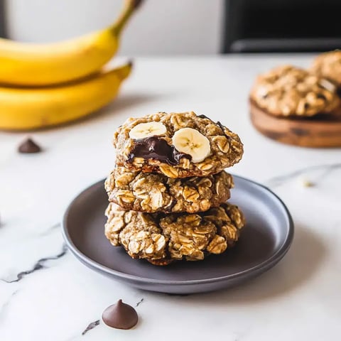 A stack of three oatmeal cookies with chocolate and banana slices on a gray plate, with bananas and chocolate chips in the background.