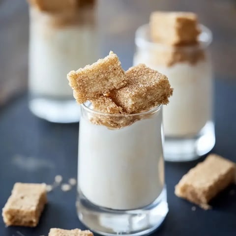 Three glasses of creamy dessert topped with brown sugar cubes are displayed on a dark surface.