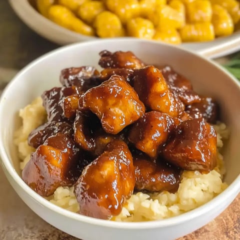 A bowl of rice topped with succulent pieces of glazed chicken in a rich brown sauce, with a side of yellow corn in the background.